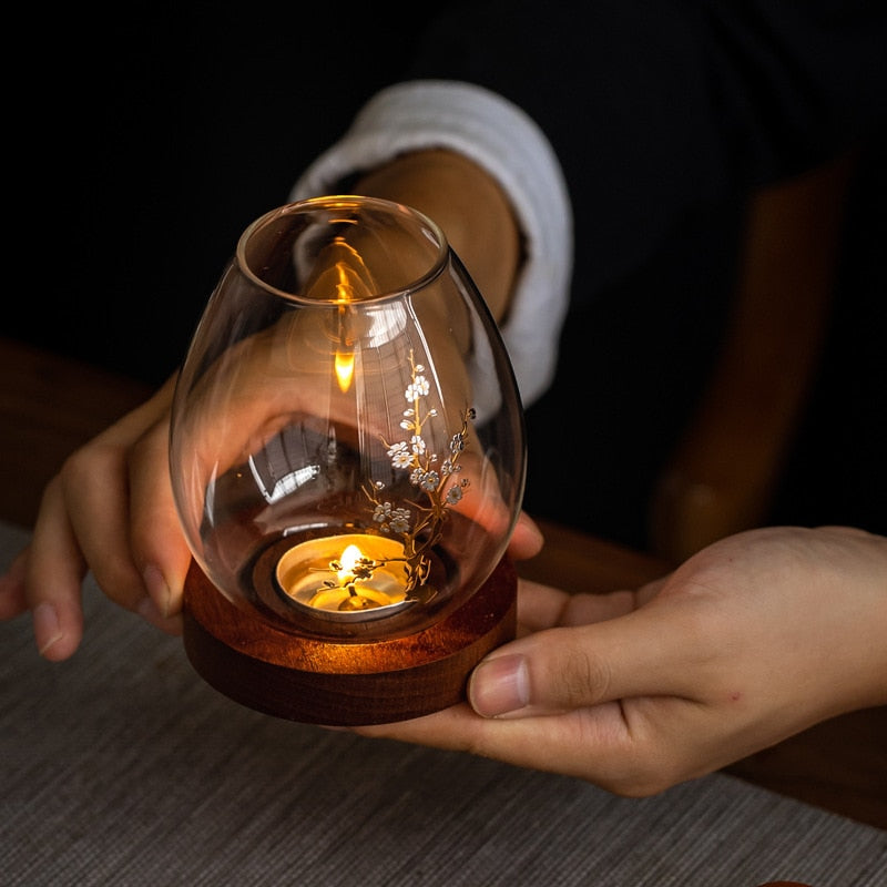 Person holding glass candle holder with blossom branch and flowers on timber base